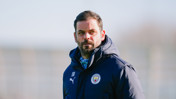 A coach wearing a Manchester City jacket stands outdoors. The jacket features the club logo and Puma branding.