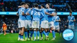A Manchester City football team huddle on the field, players in light blue jerseys with blurred faces, celebrating a goal at a crowded stadium.