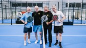 Four men holding padel rackets pose on a blue padel court.