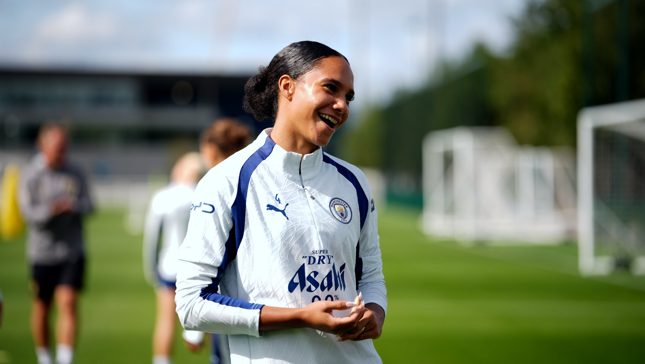 A woman in a Manchester City training outfit is smiling on a football field during a training session.