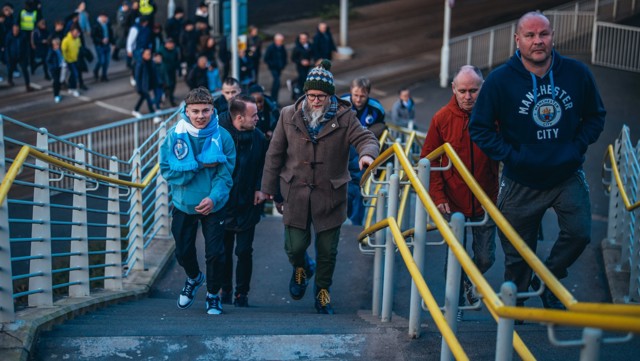 A group of Manchester City fans, including children and adults, are seen climbing stairs. They are dressed in Manchester City apparel, including scarves and sweatshirts. Other fans are walking in the background. A group of Manchester City fans, including children and adults, are seen climbing stairs. They are dressed in Manchester City apparel, including scarves and sweatshirts. Other fans are walking in the background.
