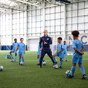 A group of young football players in Manchester City attire participate in a training session indoors with a coach guiding them.