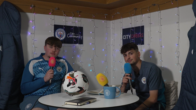 Two men holding microphones participate in a discussion. Manchester City logos are visible behind them, along with fairy lights.