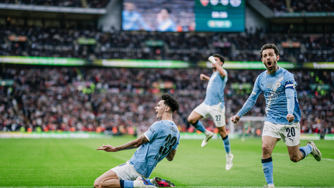Three soccer players in light blue jerseys celebrating a goal on the field with a scoreboard displaying the score in the background.