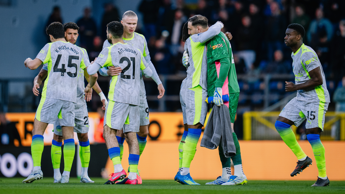 Soccer players in grey and neon kits celebrating together on the field following a match. The players are hugging and wearing uniforms with various numbers and names visible.