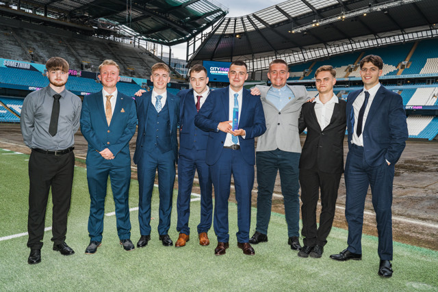 A group of men dressed in suits standing on the grass field at Etihad Stadium. The stadium is partially visible in the background, with empty stands.