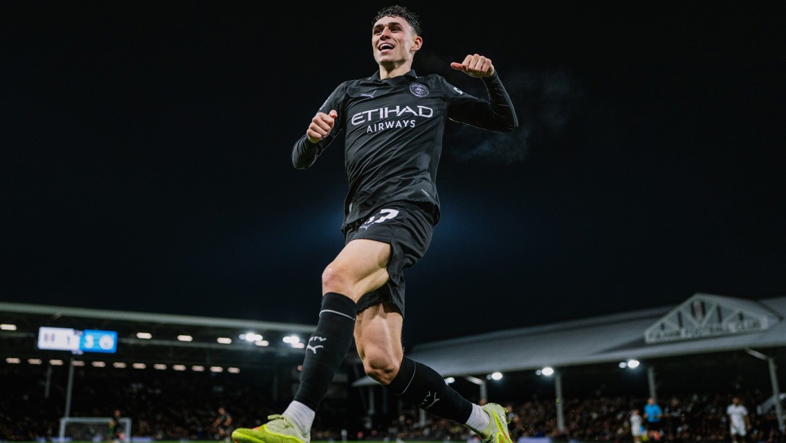 Manchester City player wearing a black kit, celebrating a goal with Etihad Airways sponsorship visible. The player is mid-jump. The stadium in the background includes the scoreboard displaying 5 - 1.