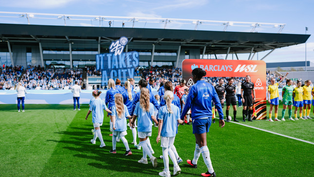 Football players and mascots walk onto the pitch at a Manchester City stadium with a large 'It Takes A City' banner. Teams line up for the WSL match under clear skies.