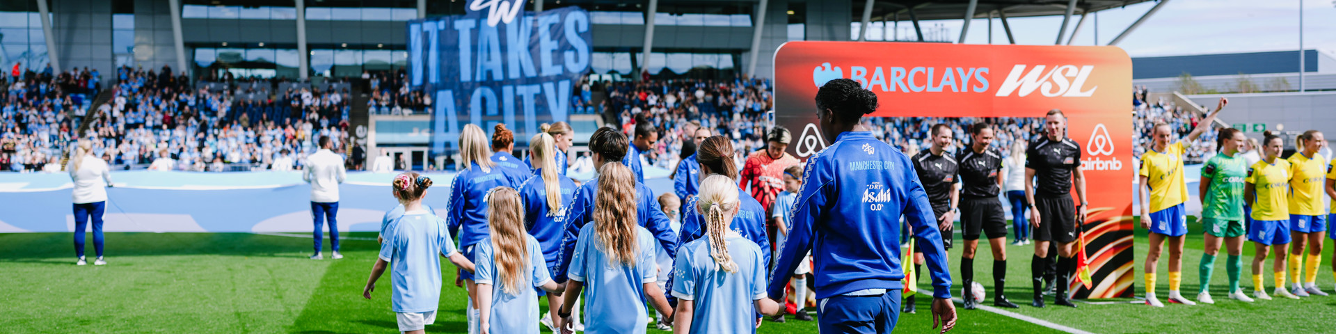 Football players and mascots walk onto the pitch at a Manchester City stadium with a large 'It Takes A City' banner. Teams line up for the WSL match under clear skies.