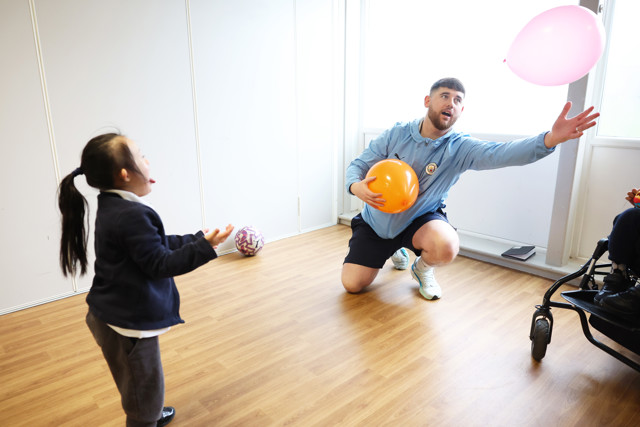 An adult kneels on the floor holding a balloon, interacting with a child reaching for another balloon in a room with wooden flooring.