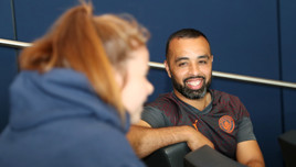Two individuals are conversing indoors. One person is wearing a gray sports shirt with the Manchester City logo.