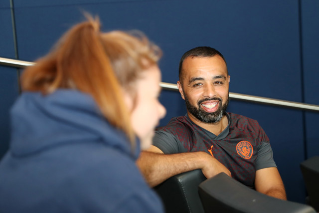 Two individuals are conversing indoors. One person is wearing a gray sports shirt with the Manchester City logo.