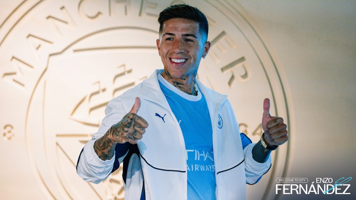 Person in a Manchester City jacket giving two thumbs up. Background features Manchester City crest and text 'Welcome to City Enzo Fernandez'.