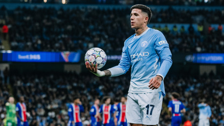 A football player in a Manchester City kit holding a ball during a match with a crowded stadium in the background.