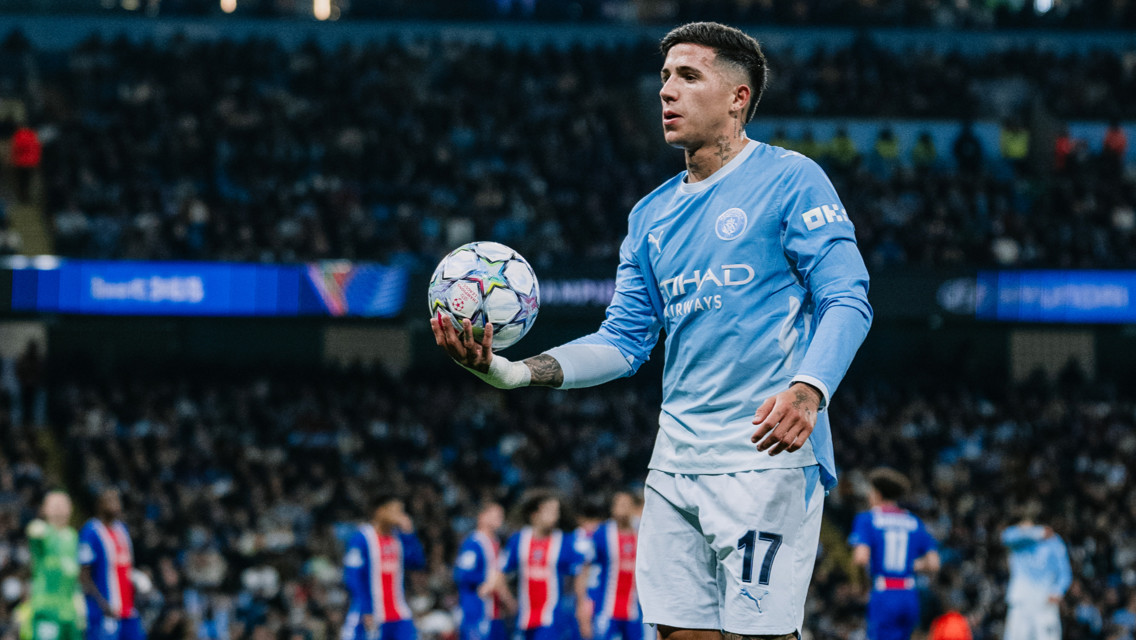 A football player in a Manchester City kit holding a ball during a match with a crowded stadium in the background.