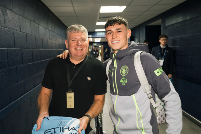 Two individuals standing in a corridor with one holding a signed Manchester City jersey.