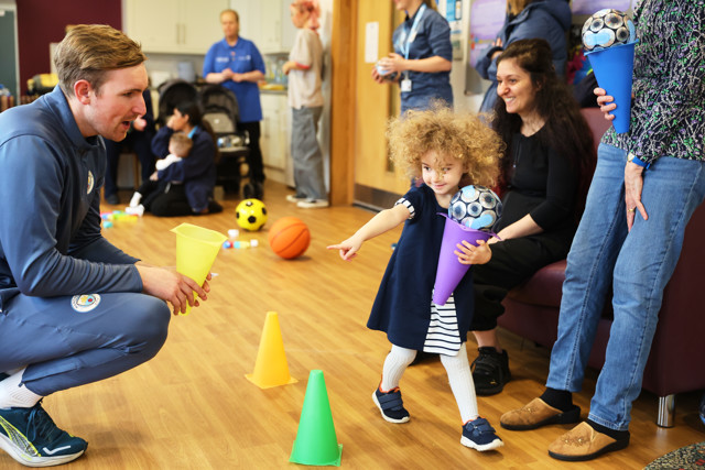 A community activity involving children and adults at a venue. A child holds a toy football in a cone, while a person in a Manchester City tracksuit engages with them. Various sports balls are scattered around the room.