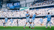 Manchester City players celebrate on the field at the Etihad Stadium during a Premier League match, with a large crowd and scoreboard showing behind them.