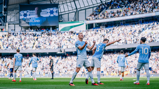 Manchester City players celebrate on the field at the Etihad Stadium during a Premier League match, with a large crowd and scoreboard showing behind them.
