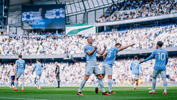 Manchester City players celebrate on the field at the Etihad Stadium during a Premier League match, with a large crowd and scoreboard showing behind them.