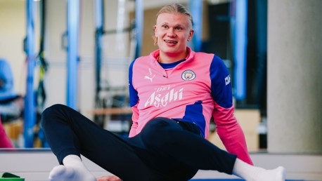 A person wearing a Manchester City training tracksuit in pink and blue is sitting on the floor with one leg extended.