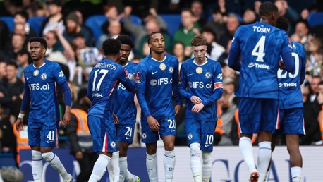 Group of Chelsea FC players in blue jerseys, interacting on the field during a match with a crowd in the background.