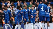 Group of Chelsea FC players in blue jerseys, interacting on the field during a match with a crowd in the background.