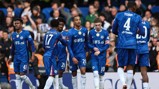 Group of Chelsea FC players in blue jerseys, interacting on the field during a match with a crowd in the background.