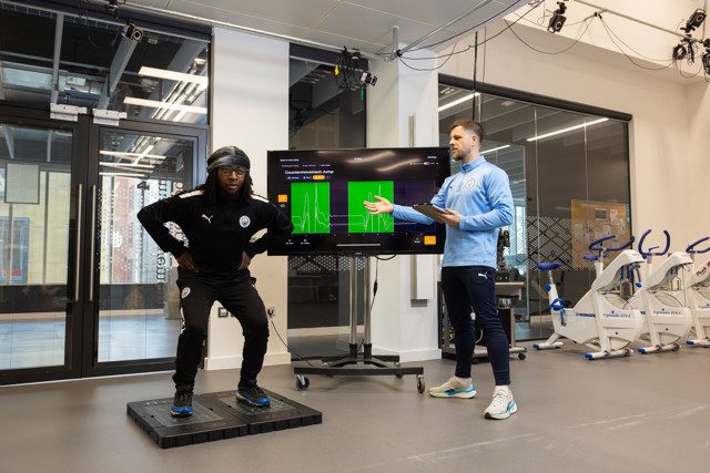 Two individuals in Manchester City tracksuits are conducting a training session with sports equipment, analyzing data on a large screen.