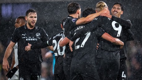 Group of Manchester City players in black kits celebrating a goal, hugging in the rain during a match, with Premier League badges visible on their shirts.