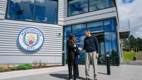 Two people shaking hands outside a modern building with the Manchester City logo on the wall.