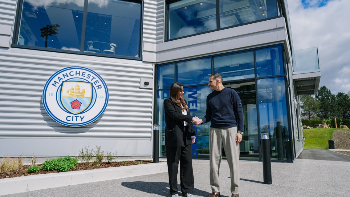 Two people shaking hands outside a modern building with the Manchester City logo on the wall.