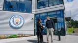 Two people shaking hands outside a modern building with the Manchester City logo on the wall.