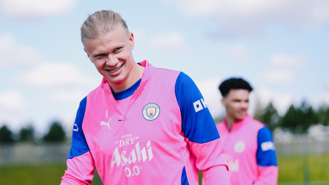 Two individuals wearing Manchester City training kit, featuring pink and blue colors, are outdoors during a training session.