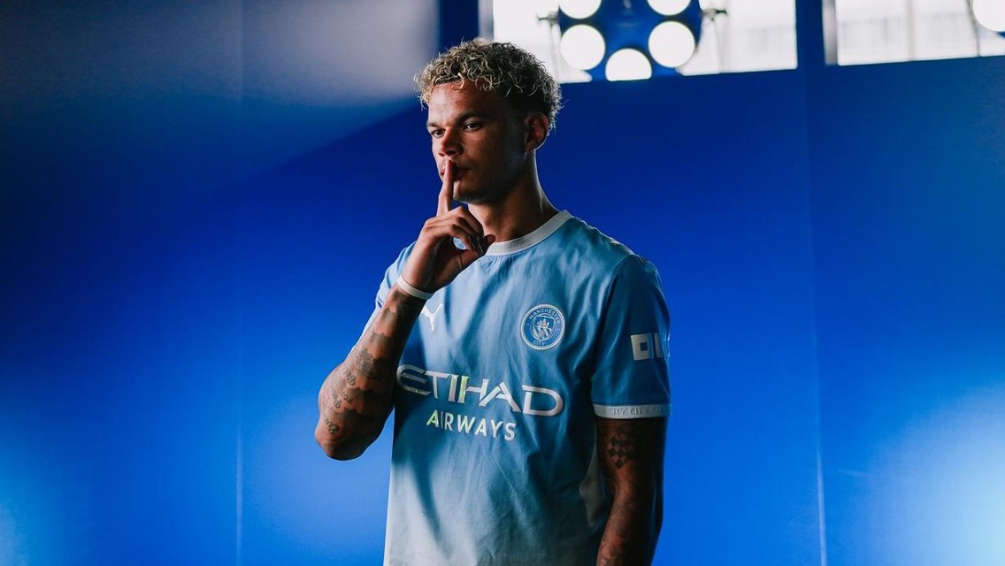 A football player wearing a Manchester City jersey poses against a blue background, with his finger over his lips in a gesture.