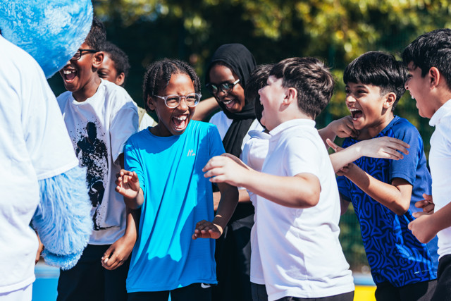 A group of children with their faces blurred out are gathered around a mascot dressed in a blue furry costume.