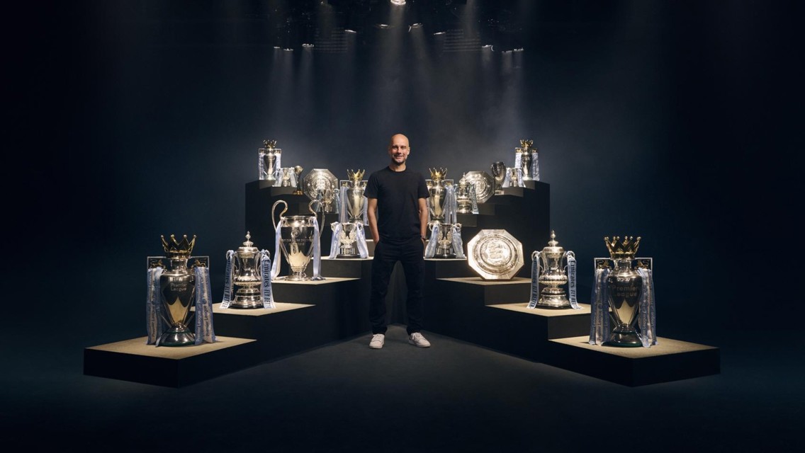 A person standing among various football trophies on platforms with dramatic lighting.