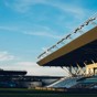 An empty football stadium with blue seats and a modern roof structure under a clear blue sky. The pitch is visible, and the lighting fixtures are mounted on the roof.