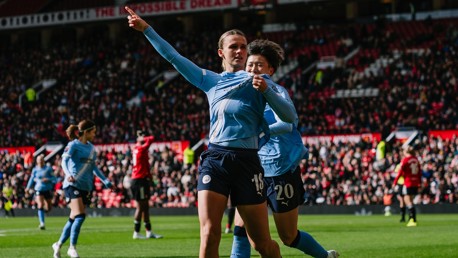 Players in blue jerseys celebrating on the field during a match, with one pointing upwards. A crowd in the stadium watches the action.