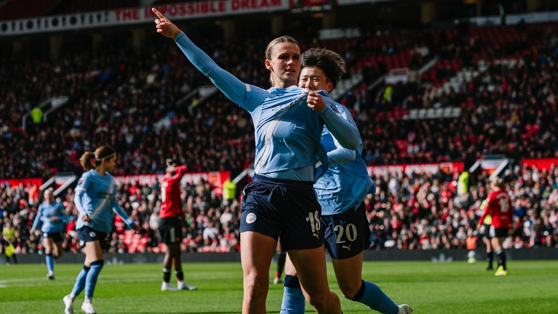 Players in blue jerseys celebrating on the field during a match, with one pointing upwards. A crowd in the stadium watches the action.