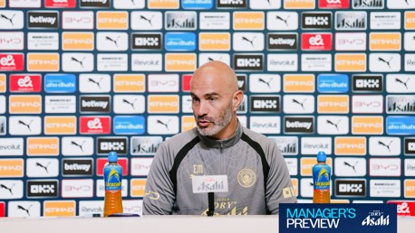 A man at a Manchester City press conference sits in front of a wall filled with various sponsor logos.