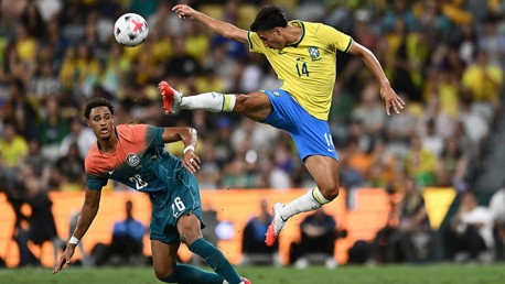 A soccer player in a yellow jersey jumps to kick the ball while another player in a multicolored jersey tries to defend during a match.