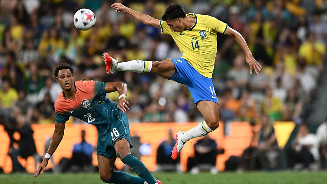 A soccer player in a yellow jersey jumps to kick the ball while another player in a multicolored jersey tries to defend during a match.
