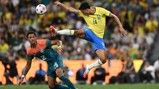 A soccer player in a yellow jersey jumps to kick the ball while another player in a multicolored jersey tries to defend during a match.