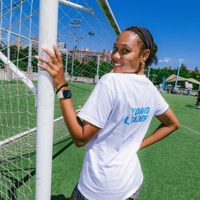 A person wearing a white t-shirt with 'YOUNG LEADER' printed on the back, standing by a soccer goal on a field.