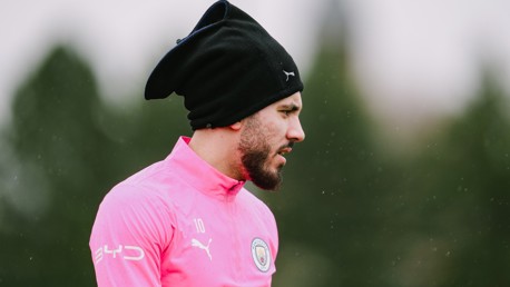 Individual wearing a pink Manchester City training jacket and a black hood or beanie, with blurry face, outdoors.