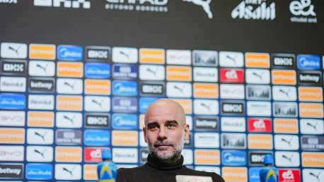 A press conference setup with a blurred individual seated in front of a backdrop displaying various sponsor logos, including Puma and Etihad.