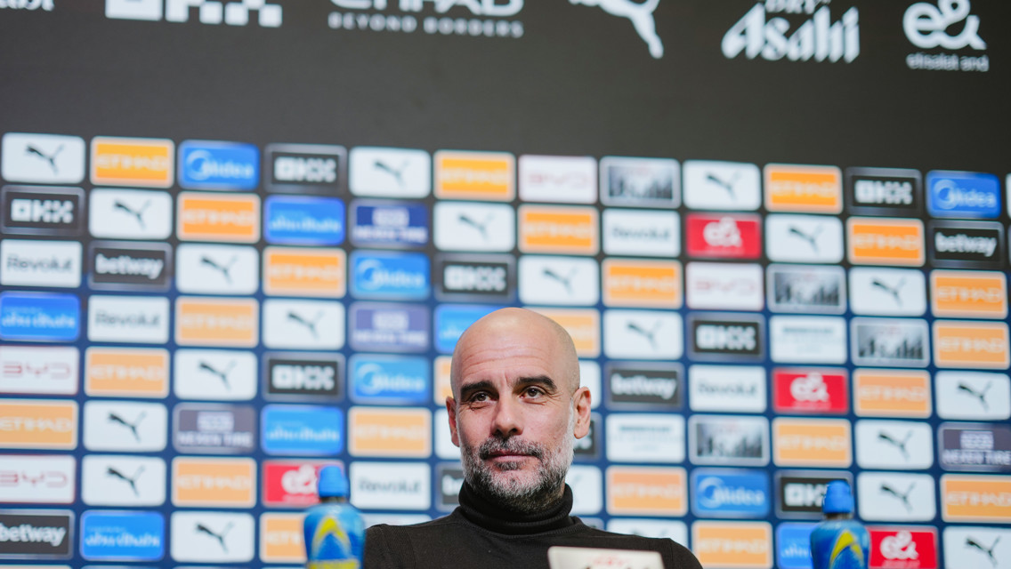 A press conference setup with a blurred individual seated in front of a backdrop displaying various sponsor logos, including Puma and Etihad.