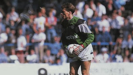 A football goalkeeper in a green kit prepares to kick the ball during a match, with a crowd in the background.