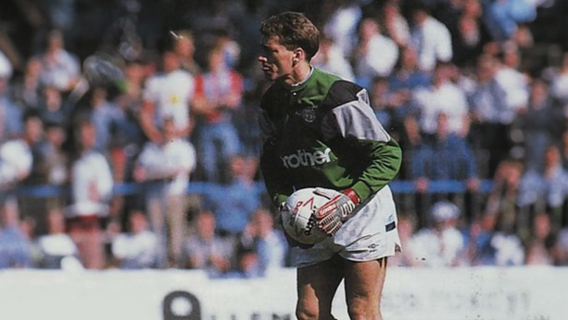 A football goalkeeper in a green kit prepares to kick the ball during a match, with a crowd in the background.